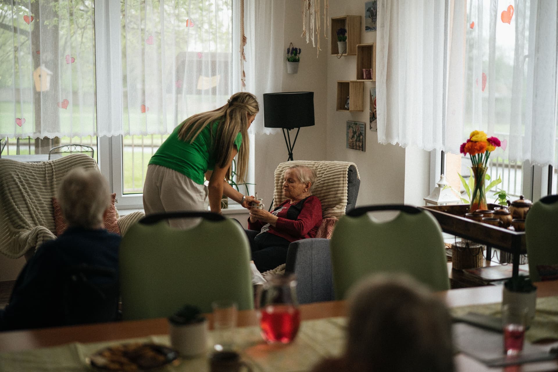A caregiver handing a drink to an elderly woman seated in a comfortable home